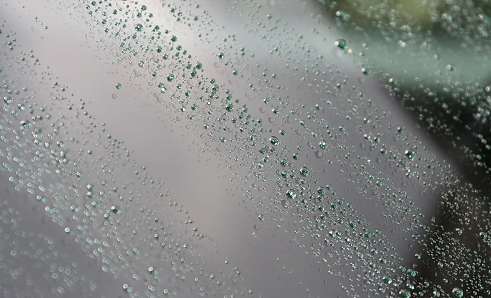 Viele kleine Wassertropfen auf einer grauen Glasfläche, diagonal angeordnet, mit unscharfem grünlichem Hintergrund rechts oben.