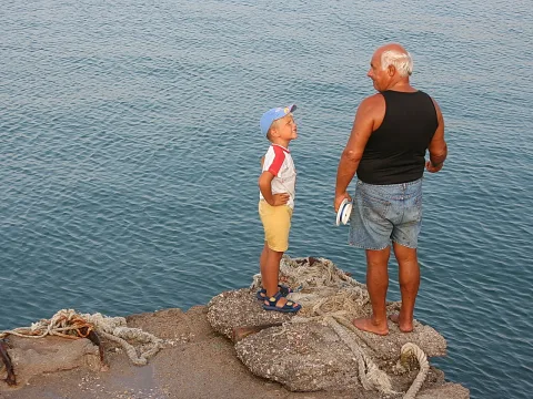 Ein älterer Mann mit Shorts und schwarzem Tanktop steht barfuß auf einer Hafenmole und blickt zu einem lachenden Jungen mit blauer Kappe, neben ihnen liegen dicke Taue auf dem Stein, im Hintergrund ruhiges Meer.