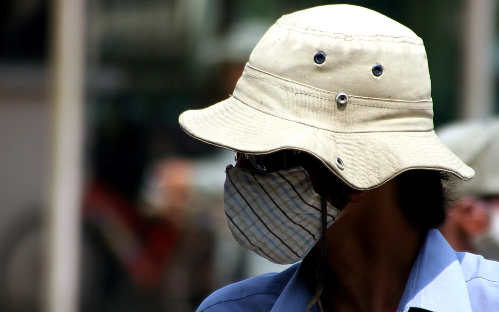 Ein Mensch von der Seite mit beigem Buckelhut und karierter Stoffmaske vor Nase und Mund, im Hintergrund unscharfe Straße.