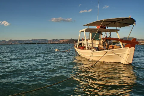 Weißes Fischerboot mit braunem Bug und blauem Sonnendach liegt vertäut auf ruhigem, blaugrünem Meer. Im Hintergrund erheben sich braune Hügel unter klarem Himmel, Abendlicht fällt warm auf das Wasser.