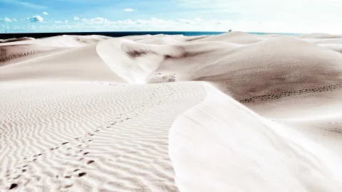 Helle Sanddünen mit geschwungenen Rippeln und Fußspuren, im Hintergrund das blaue Meer unter einem Himmel mit Wolken. Auf einer Düne stehen zwei winzige Personen.