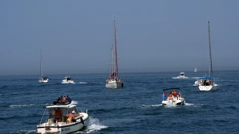 Mehrere Segel- und Motorboote fahren auf blauem Meer, im Vordergrund ein weißes Motorboot mit Menschen an Bord, dahinter ein Segelboot mit roten Masten unter diesigem Himmel.