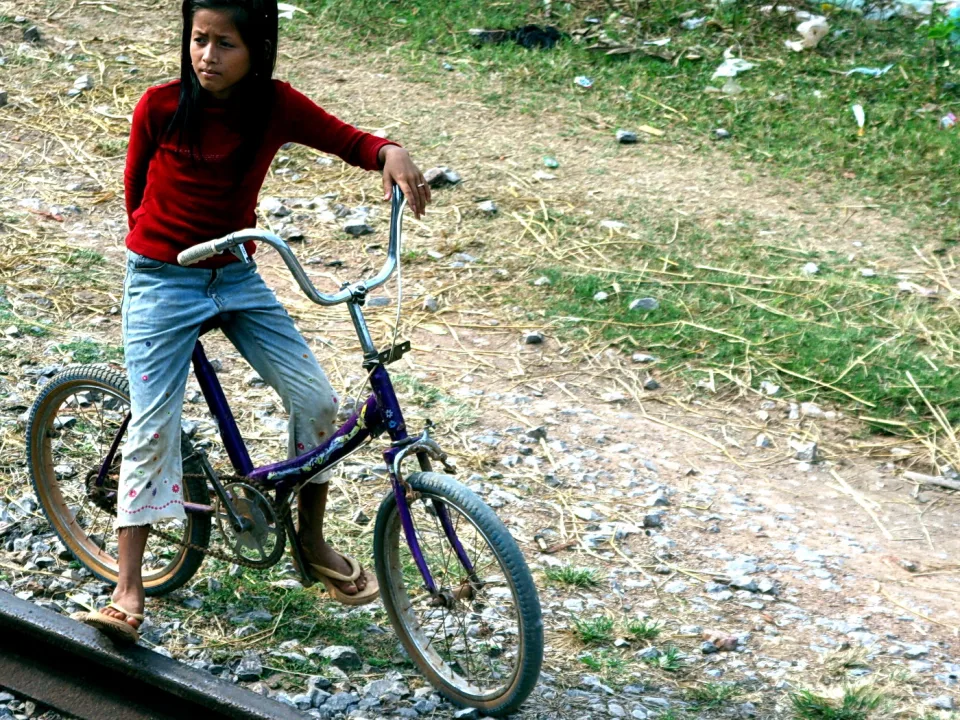 Ein Mädchen mit langen dunklen Haaren und rotem Pullover steht auf einem lila Fahrrad auf einer Schotterfläche neben Bahngleisen, im Hintergrund Gras und verstreuter Müll.