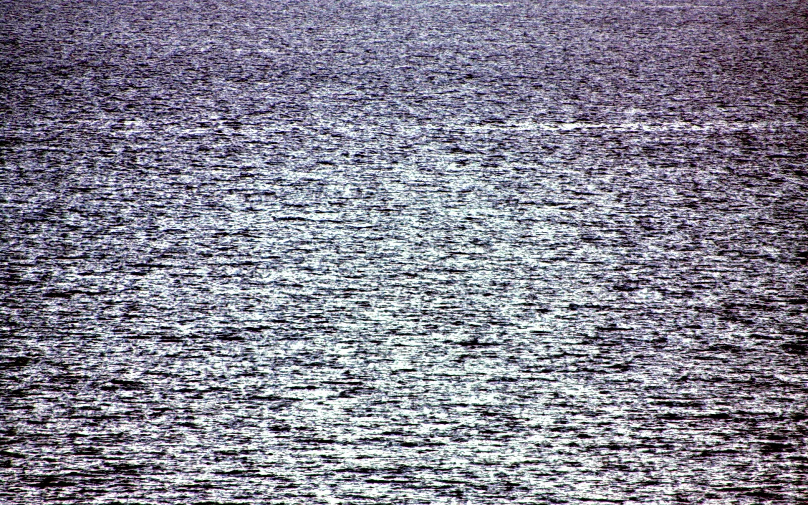 Wasseroberfläche eines Meeres, auf der sich helles Sonnenlicht in vielen kleinen Reflexen bricht. Die Fläche wirkt grau-blau bis violett.