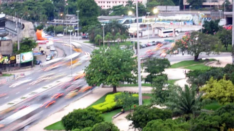 Langzeitbelichtung einer mehrspurigen Straße in Rio de Janeiro bei Nacht, Autos erzeugen rote und weiße Lichtstreifen, rechts grüne Bäume und Palmen in einem Stadtpark.