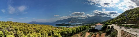 Panoramablick über bewaldete Hügel auf eine blaue Bucht mit Bergen im Hintergrund, rechts ein weißes Gebäude mit rotem Ziegeldach und eine Straße.