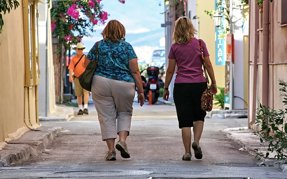 Zwei Frauen von hinten in Sommerkleidung gehen eine schmale Gasse entlang, links eine gelbe Hauswand, rechts ein Roller, im Hintergrund ein Mann mit Hut und orangefarbenem Hemd.