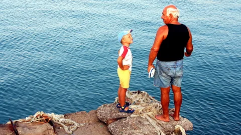 Ein älterer kahlköpfiger Mann in schwarzem Tanktop und Jeansshorts steht barfuß auf einem Felsen am Meer. Ihm gegenüber steht ein kleiner Junge mit blauer Kappe, gelben Shorts und Sandalen, der zu ihm aufschaut. Im Vordergrund liegen Seile auf dem Stein.