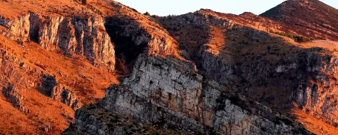 Steile Felswände aus geschichtetem hellem Kalkstein und rotbraunen Hängen, von tief stehender Abendsonne orange angestrahlt.
