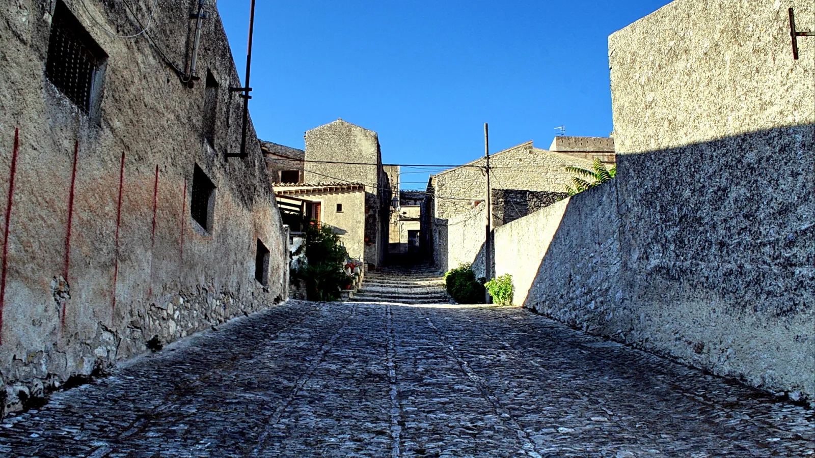 Schmale, gepflasterte Gasse zwischen alten Steinmauern unter klarem blauem Himmel, am Ende steigt der Weg leicht an.