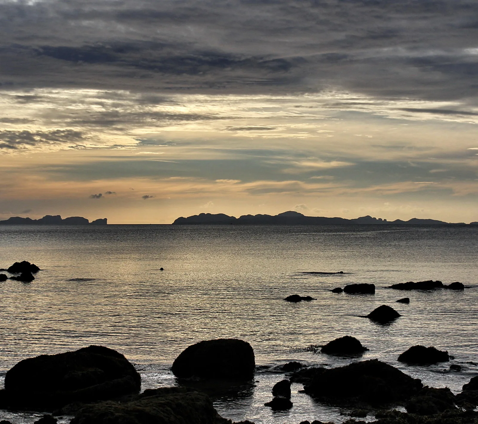 Meer bei Abendstimmung mit dunklen Felsen im Vordergrund und langgezogenen Inseln am Horizont unter einem bewölkten Himmel