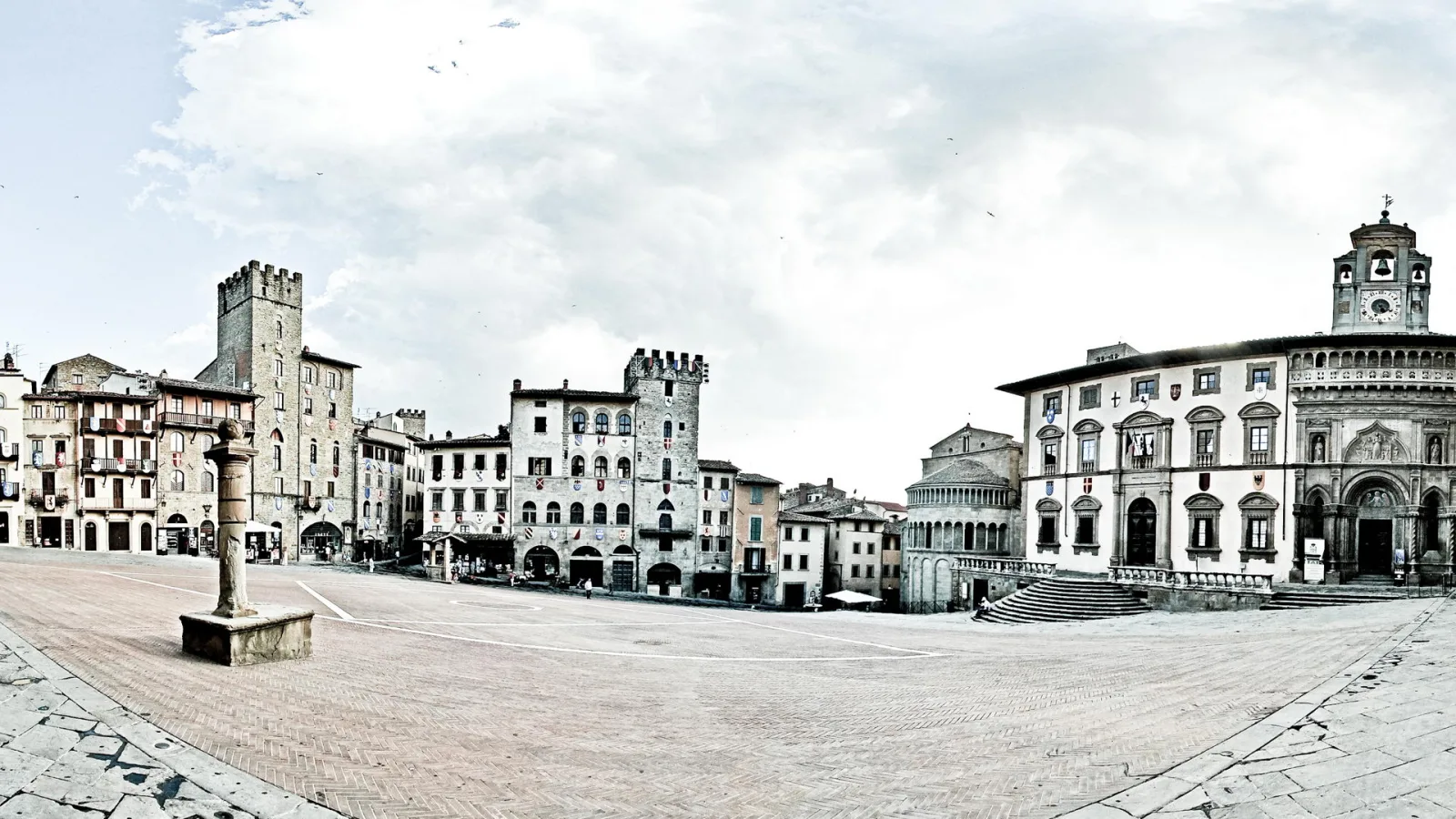 Weiter Rathausplatz mit Kopfsteinpflaster, links mittelalterliche Häuser und ein Turm, rechts ein Palast mit Glockenturm und Freitreppe, darüber bewölkter Himmel.