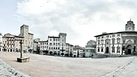Weiter Rathausplatz mit Kopfsteinpflaster, links mittelalterliche Häuser und ein Turm, rechts ein Palast mit Glockenturm und Freitreppe, darüber bewölkter Himmel.