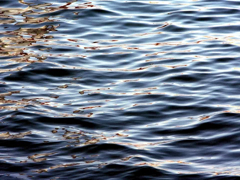 Nahaufnahme einer bewegten Wasseroberfläche mit dunkelblauen Wellen und goldfarbenen Lichtreflexen, besonders links oben.