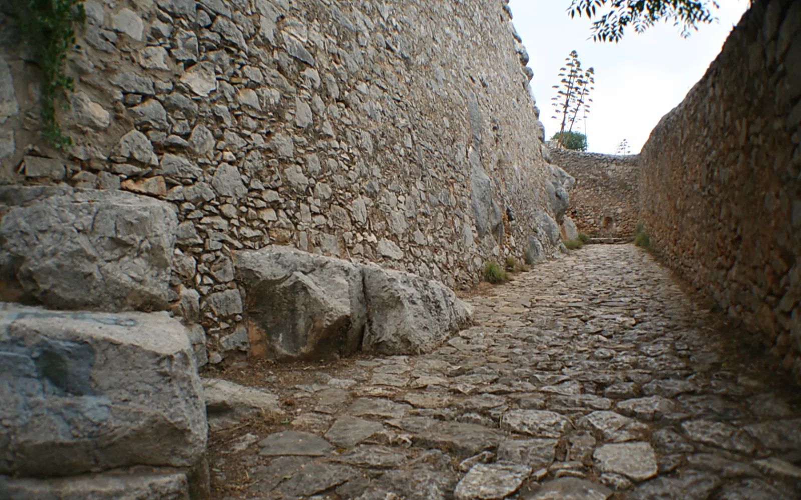 Kopfsteinpflasterte Gasse zwischen hohen Bruchsteinmauern, links ein großer Felsbrocken, am Ende der Gasse ein Baum vor hellem Himmel.