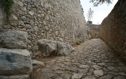 Kopfsteinpflasterte Gasse zwischen hohen Bruchsteinmauern, links ein großer Felsbrocken, am Ende der Gasse ein Baum vor hellem Himmel.