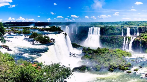 Panorama der Iguazú-Wasserfälle mit mehreren weißen Kaskaden, dichtem grünen Dschungel und Felseninseln unter blauem Himmel mit Wolken.
