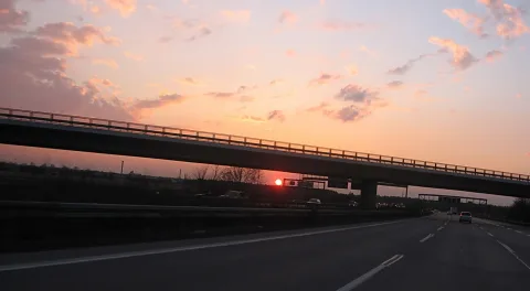 Autobahn mit Fahrbahnmarkierungen führt unter einer Brücke hindurch, im Hintergrund geht die rote Sonne hinter Bäumen unter, der Himmel ist rosa und orange mit Wolken.
