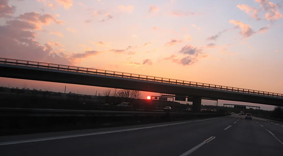 Autobahn mit Fahrbahnmarkierungen führt unter einer Brücke hindurch, im Hintergrund geht die rote Sonne hinter Bäumen unter, der Himmel ist rosa und orange mit Wolken.