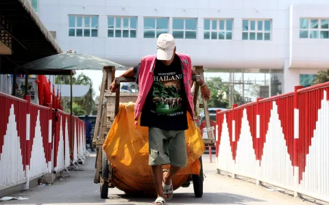 Ein Mann mit Basecap und dunkelrotem Shirt schiebt einen Holzkarren mit orangefarbener Plane über eine Brücke mit rot-weißem Geländer.