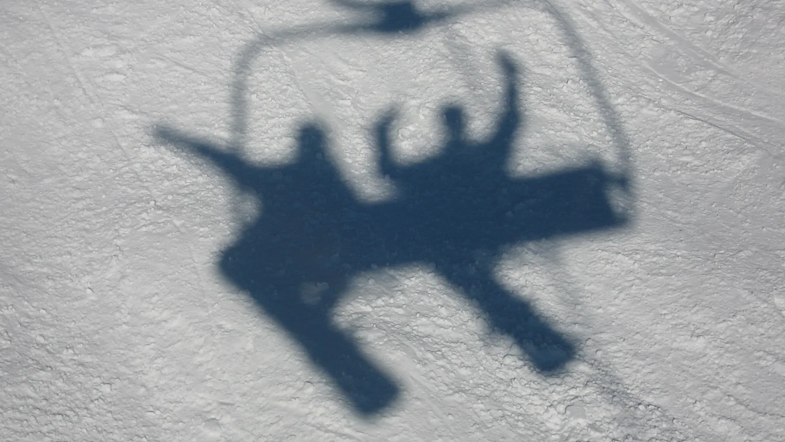 Schatten von zwei Personen auf einem Sessellift über weißem Schnee, Arme nach oben gestreckt, Skier nach unten hängend.