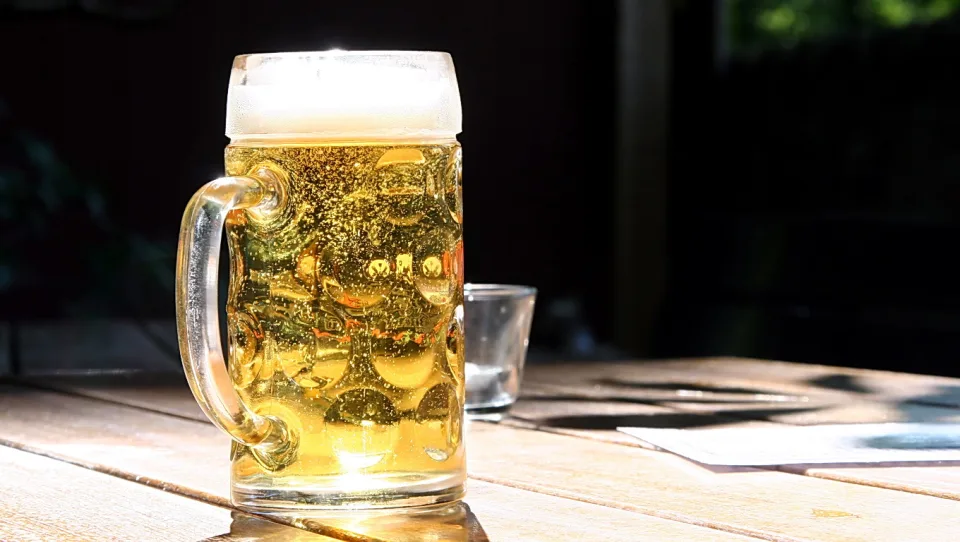 Ein Henkelkrug mit hellem Bier und Schaumkrone steht im Sonnenlicht auf einem Holztisch, daneben ein leeres Glas, vor dunklem Hintergrund.