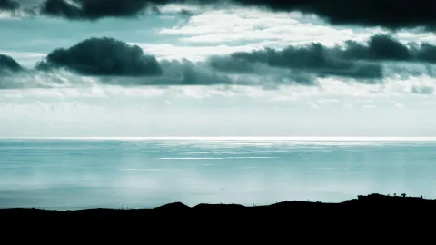 Ruhige, graublaue Meeresfläche unter dunklen Wolkenbänken, am unteren Bildrand eine dunkle Hügelkette
