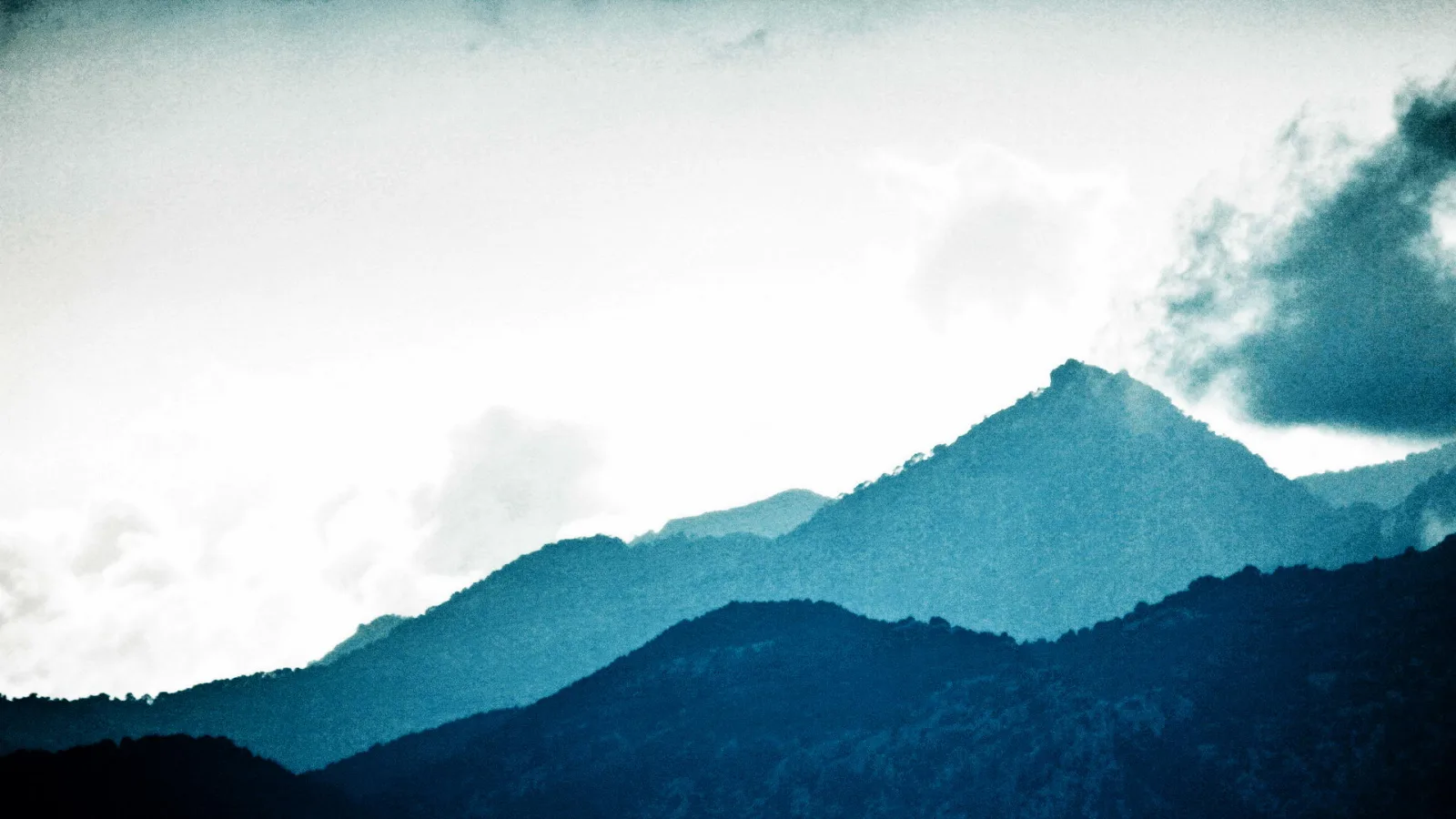 Mehrere blau getönte Bergketten hintereinander, der hellste Gipfel rechts der Mitte, darüber heller Himmel mit Wolkenfetzen.