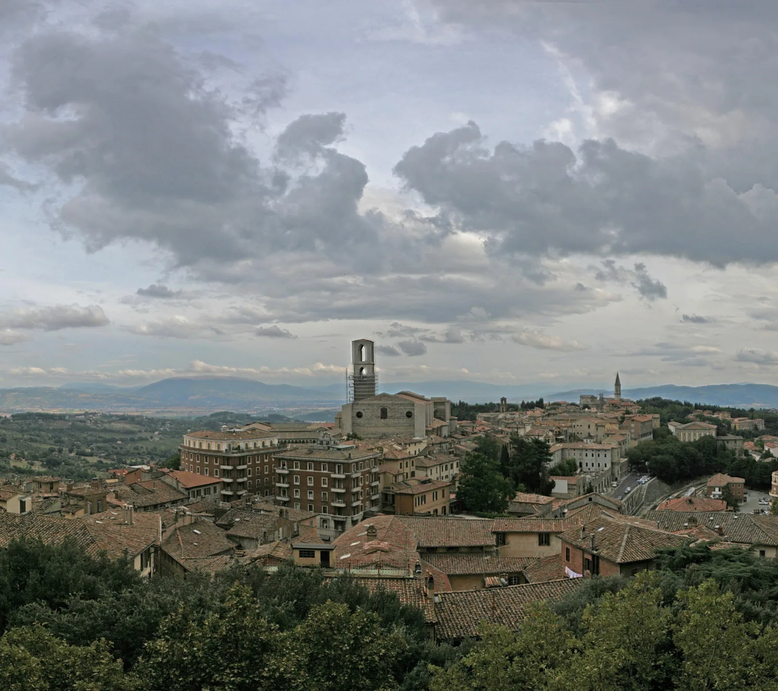 Panoramablick auf eine hügelige italienische Altstadt mit dicht stehenden Ziegeldächern, einem Kirchturm mit Gerüst in der Mitte und bewaldeten Hängen im Vordergrund, darüber ein bewölkter Himmel.