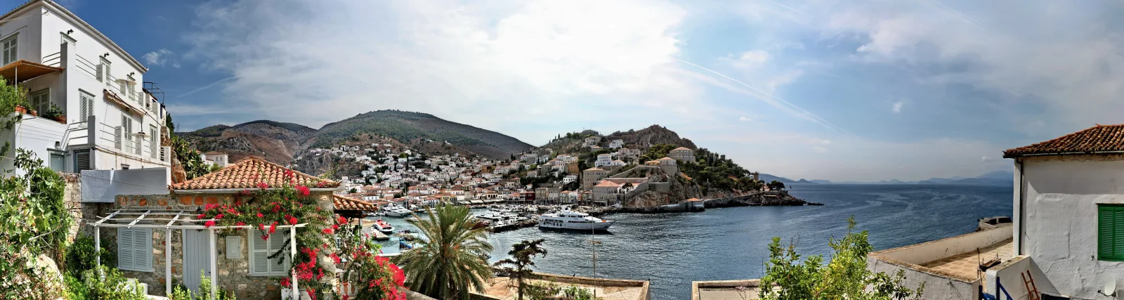 Panoramablick auf einen Hafen mit weißen Häusern an einem Hang, Booten am Kai, Palmen, Bergen und blauem Meer unter bewölktem Himmel.