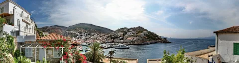 Panoramablick auf einen Hafen mit weißen Häusern an einem Hang, Booten am Kai, Palmen, Bergen und blauem Meer unter bewölktem Himmel.