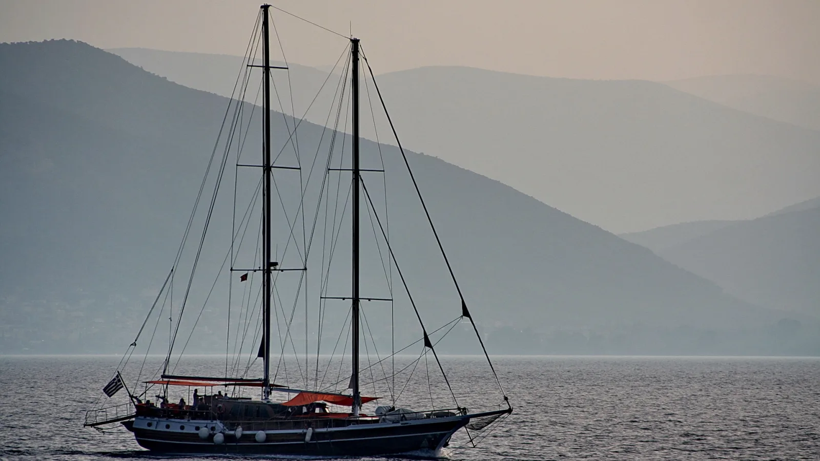 Zweimastiges Segelschiff auf ruhigem Meer vor dunstigen Bergen, am Heck eine griechische Flagge