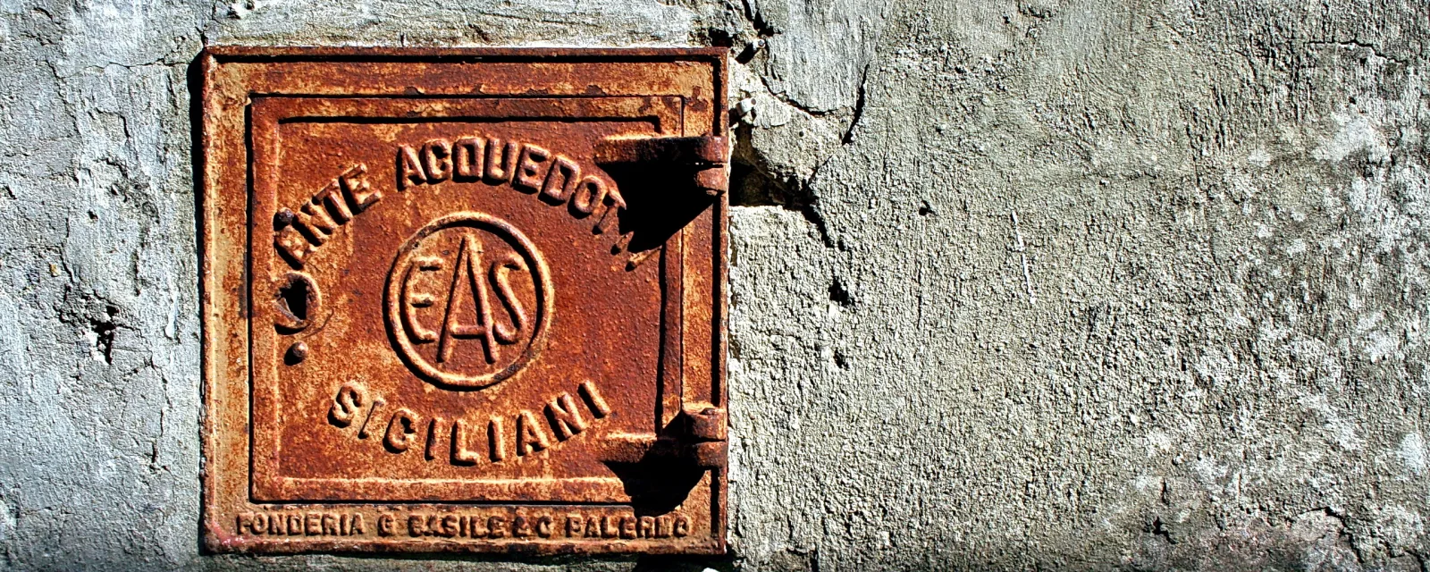 Quadratischer, rostbrauner Gussdeckel mit erhabenem Schriftzug und Logo EAS an einer verwitterten grauen Mörtelwand.