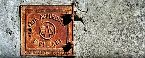 Quadratischer, rostbrauner Gussdeckel mit erhabenem Schriftzug und Logo EAS an einer verwitterten grauen Mörtelwand.