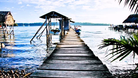 Ein langer Holzsteg führt von einem Ufer aufs ruhige blaue Wasser hinaus, links steht ein Pfahlhaus, rechts ragen Palmwedel ins Bild, im Hintergrund fährt ein Boot und ein Waldstreifen zieht sich am Horizont entlang.
