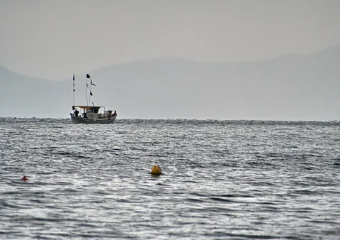 Ein kleiner Kutter mit Flaggen fährt auf einer ruhigen, grauen See, dahinter liegen im Dunst Berge am Horizont. Rechts unten schwimmt eine gelbe Boje.
