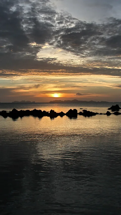 Sonnenuntergang über einem ruhigen Meer, im Vordergrund eine Reihe dunkler Felsen, darüber dichte Wolkenschichten mit orangefarbenem Lichtstreifen am Horizont.