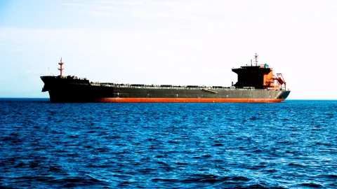 Ein dunkler Frachter mit rotem Unterwasserschiff fährt bei klarem Himmel über blaues, leicht bewegtes Meer.