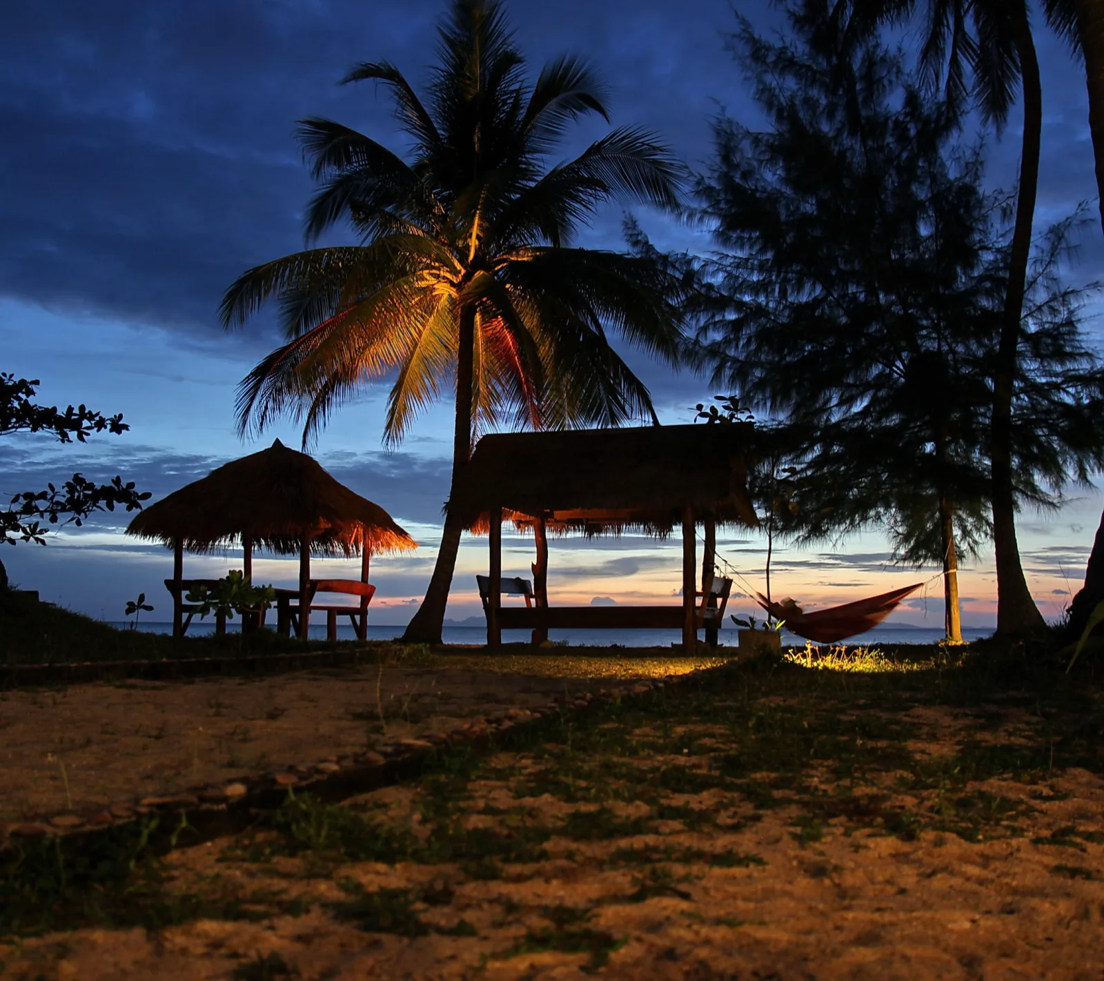 Palme, zwei strohgedeckte Unterstände und eine rote Hängematte am Strand bei Dämmerung, dahinter das Meer und ein blau-oranger Himmel