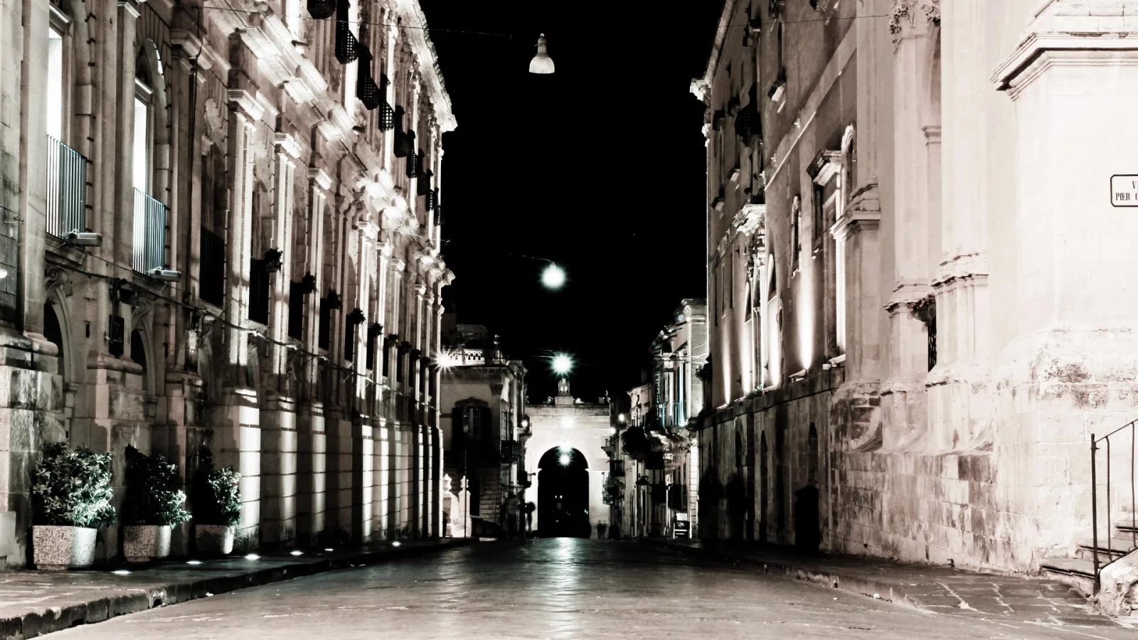 Leere Straße bei Nacht zwischen barocken Palazzi mit beleuchteten Fassaden, am Ende ein erleuchteter Torbogen.