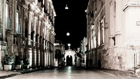 Leere Straße bei Nacht zwischen barocken Palazzi mit beleuchteten Fassaden, am Ende ein erleuchteter Torbogen.