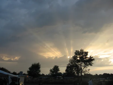 Dunkle Wolkendecke, aus der Sonnenstrahlen fächerförmig auf eine Wiese mit Bäumen und Zelten fallen. Links steht ein weißes Partyzelt, rechts eine große Baumsilhouette.