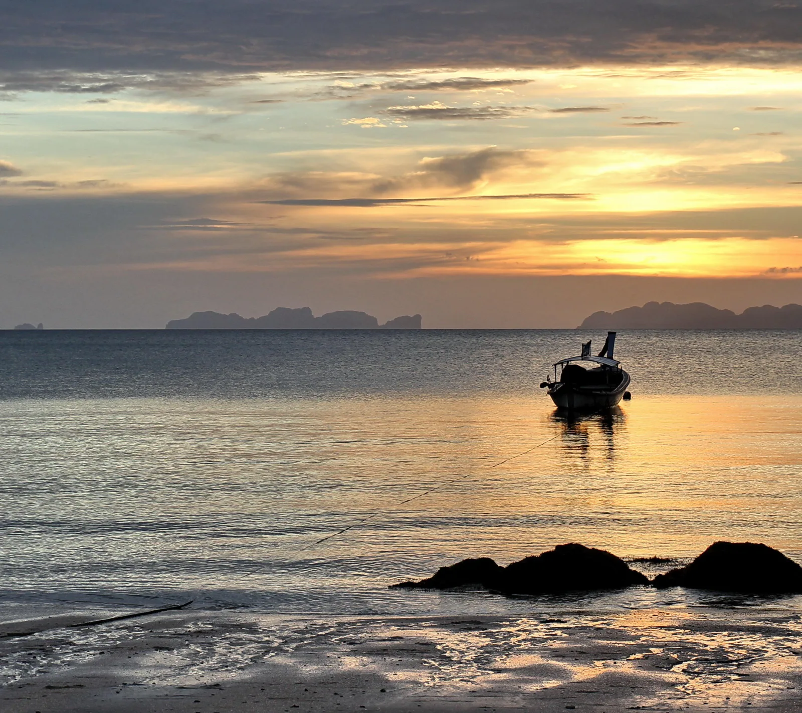 Ein kleines Boot liegt vor Anker auf ruhigem Meer, im Gegenlicht eines Sonnenuntergangs. Im Hintergrund sind Felsinseln am Horizont zu sehen, im Vordergrund dunkle Felsen am Strand.