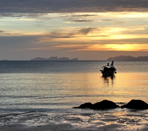 Ein kleines Boot liegt vor Anker auf ruhigem Meer, im Gegenlicht eines Sonnenuntergangs. Im Hintergrund sind Felsinseln am Horizont zu sehen, im Vordergrund dunkle Felsen am Strand.