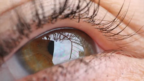 Nahaufnahme eines braun-grünen menschlichen Auges mit langen Wimpern und einer Spiegelung von Himmel und Wolken in der Iris.