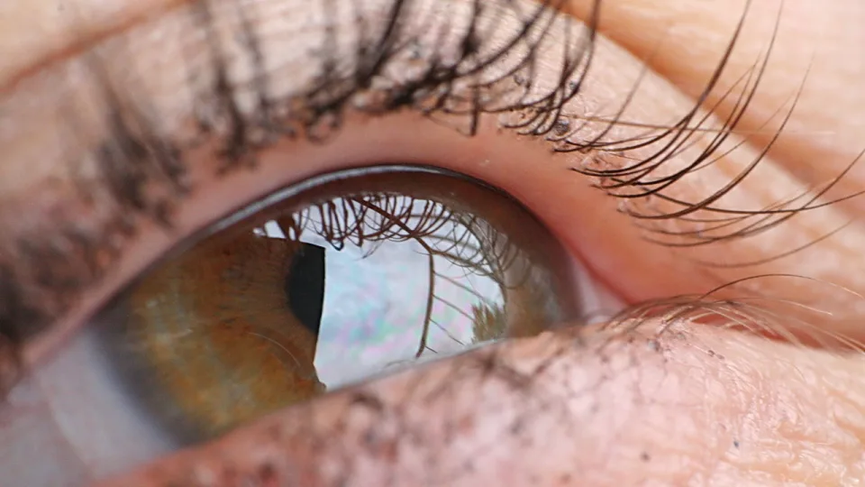 Nahaufnahme eines braun-grünen menschlichen Auges mit langen Wimpern und einer Spiegelung von Himmel und Wolken in der Iris.