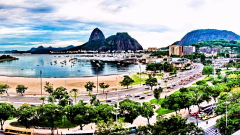 Blick über die Bucht von Botafogo in Rio de Janeiro auf den Zuckerhut, davor eine vierspurige Küstenstraße mit Autos und Bussen, Strand und Palmen im Vordergrund, dahinter Segelboote im Wasser und bewölkter Himmel.