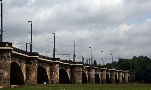 Lange Steinbrücke mit mehreren Rundbögen über eine Wiese, darüber Oberleitungen und Straßenlaternen unter bewölktem Himmel.