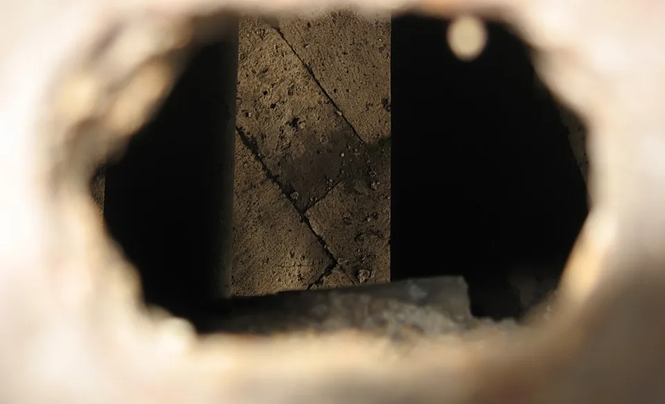 Ein runder Durchblick in einer hellen Mauer, dahinter liegt eine dunkle Öffnung mit einer grob strukturierten Steinwand.