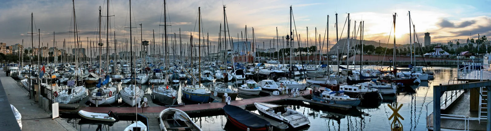 Panoramafoto eines dicht belegten Yachthafens mit zahlreichen Segelbooten und Motorbooten vor Sonnenuntergang, im Hintergrund Gebäude und ein Hügel.